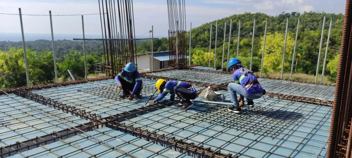 Three workers tying reinforcing steel across a suspended slab at the Cebu Technological University multi-purpose building in Borbon, with the coastline visible beyond the site.