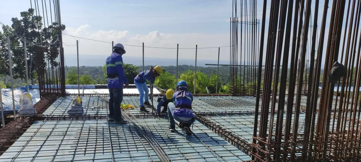 Four workers laying and tying slab reinforcement at the Cebu Technological University building in Borbon, the sea visible behind them.